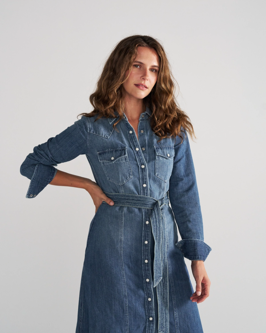 A woman wearing a long-sleeved denim shirt dress with a waist tie and button-down front.