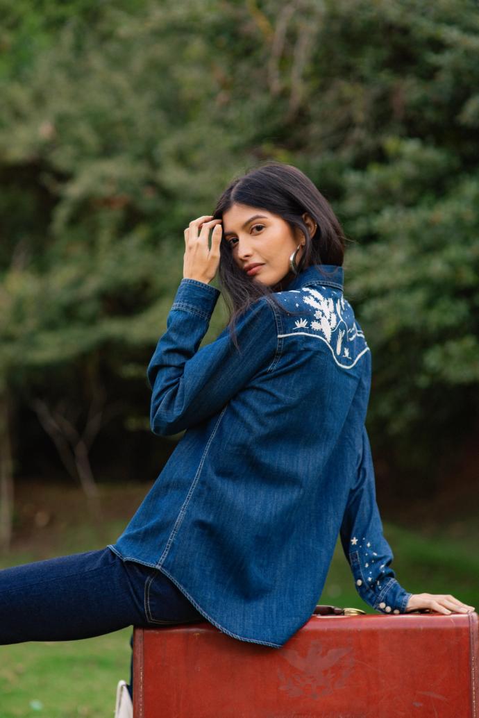 A woman in a denim shirt with floral embroidery on the back sits on a brown suitcase outdoors, with greenery in the background.