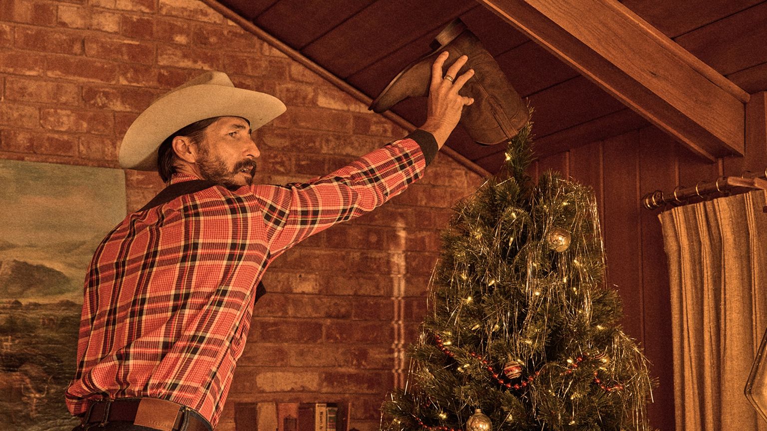 A man in a cowboy hat places a brown boot on top of a decorated Christmas tree in a wood-paneled room.