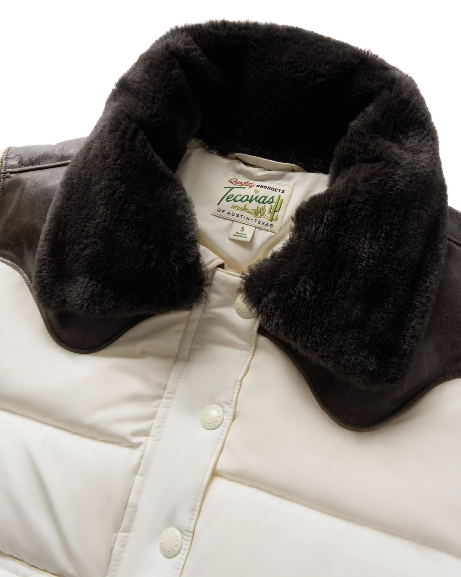 Close-up of a white puffer jacket with dark faux fur collar, brown shoulder accents, snap buttons, and a Tecovas branded label inside the neck area.