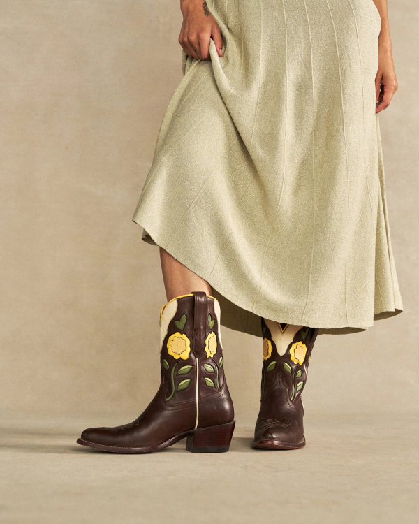 The Jolene in Truffle Cowhide. A person in a light-colored skirt displays The Jolene Cowhide brown cowboy boots decorated with yellow flower and green leaf designs, standing on a neutral backdrop.