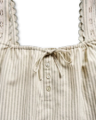 Close-up of a sleeveless striped top with lace-trimmed straps, three buttons, and a drawstring at the neckline, set against a black background.