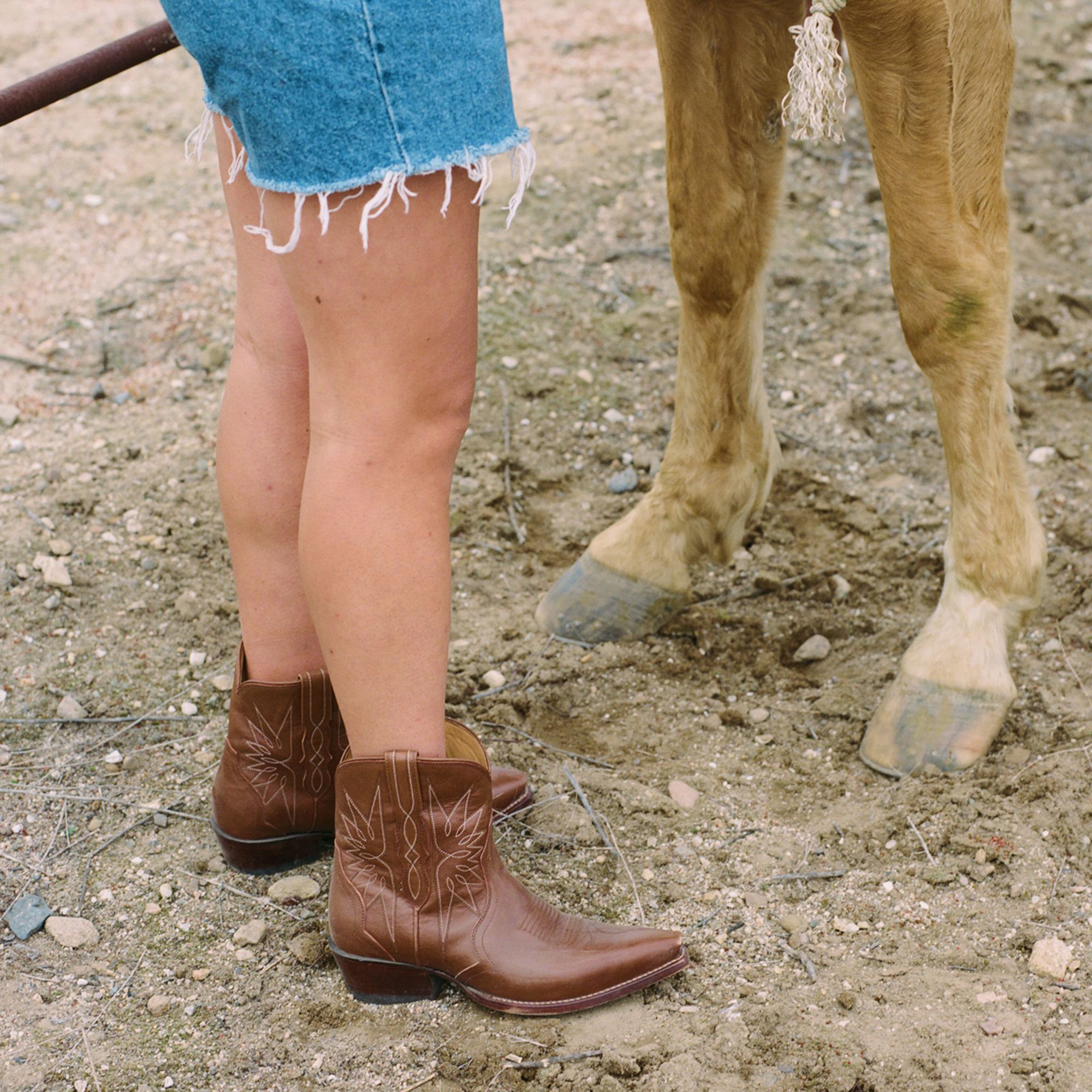 Person in brown cowboy boots and frayed denim shorts standing next to a light brown horse on a dirt ground.