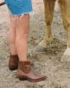 Person in brown cowboy boots and frayed denim shorts standing next to a light brown horse on a dirt ground.