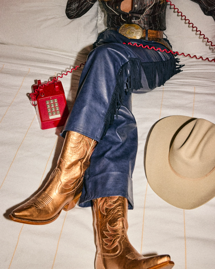 The Annie in Copper Cowhide. Person in fringed leather pants and metallic cowboy boots sitting on a bed, holding a red corded phone; The Annie Cowhide cowboy hat rests nearby.