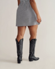 A person wearing a black and white checkered dress and black cowboy boots stands facing away, with one hand by their side.