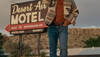 A person wearing a cowboy hat, patterned jacket, and jeans stands in front of a sign that reads “Desert Air Motel” in a rural setting.