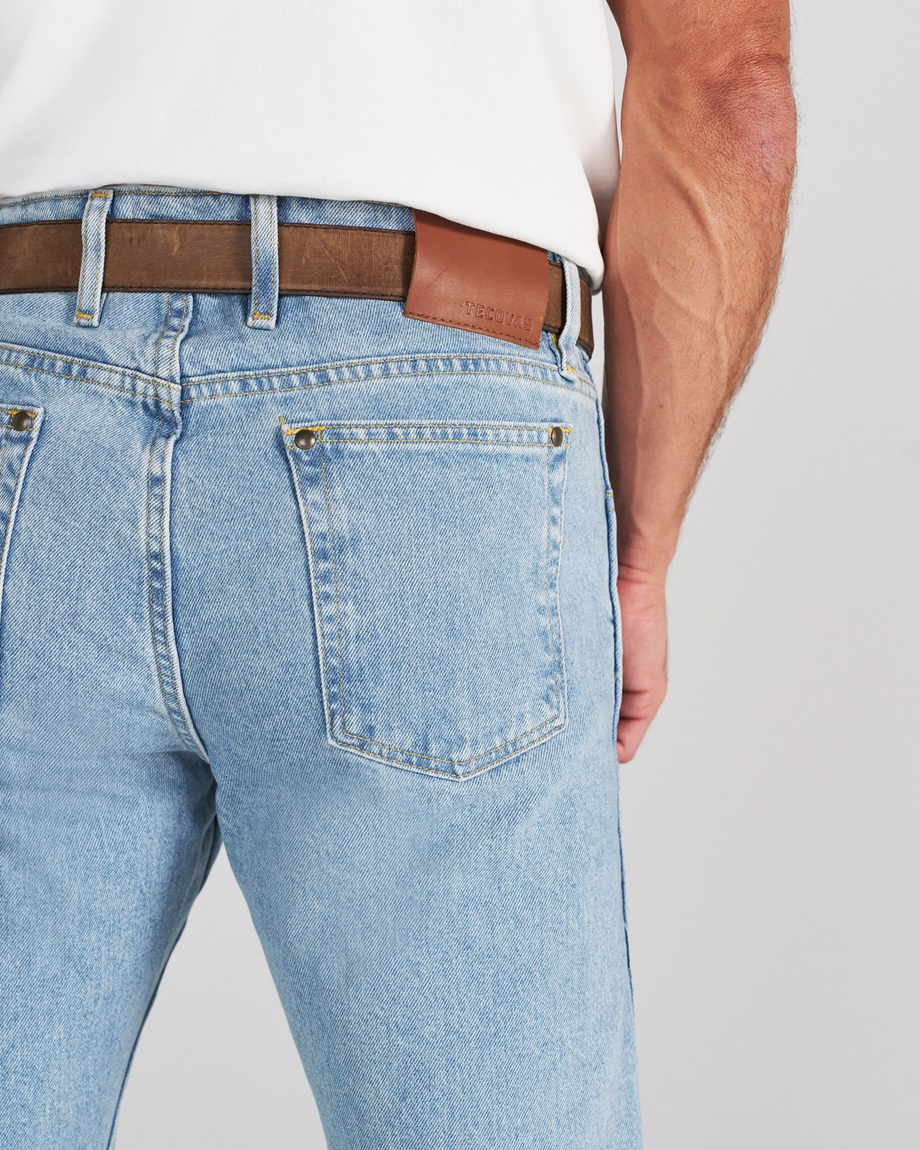 A person wearing light-wash blue jeans and a brown leather belt with a Tecovas patch on the back.