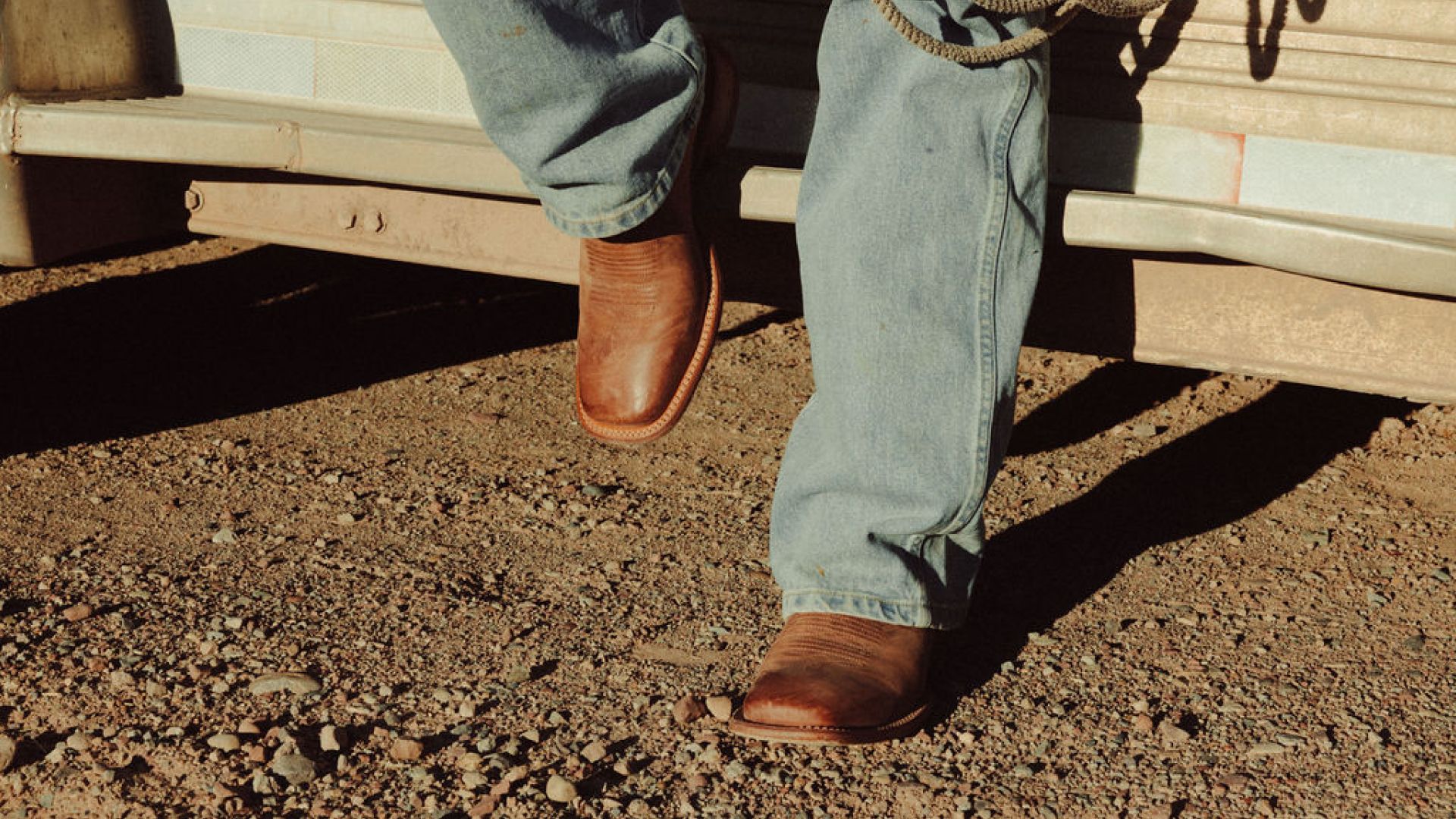 A person wearing blue jeans and brown cowboy boots steps onto gravel beside a vehicle.