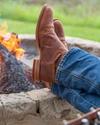 Person wearing jeans and brown boots with feet up on a stone firepit edge, relaxing by a campfire.