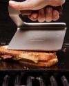 A hand presses a grilled cheese sandwich with a metal spatula on a grill, causing cheese to melt and ooze from the sides.