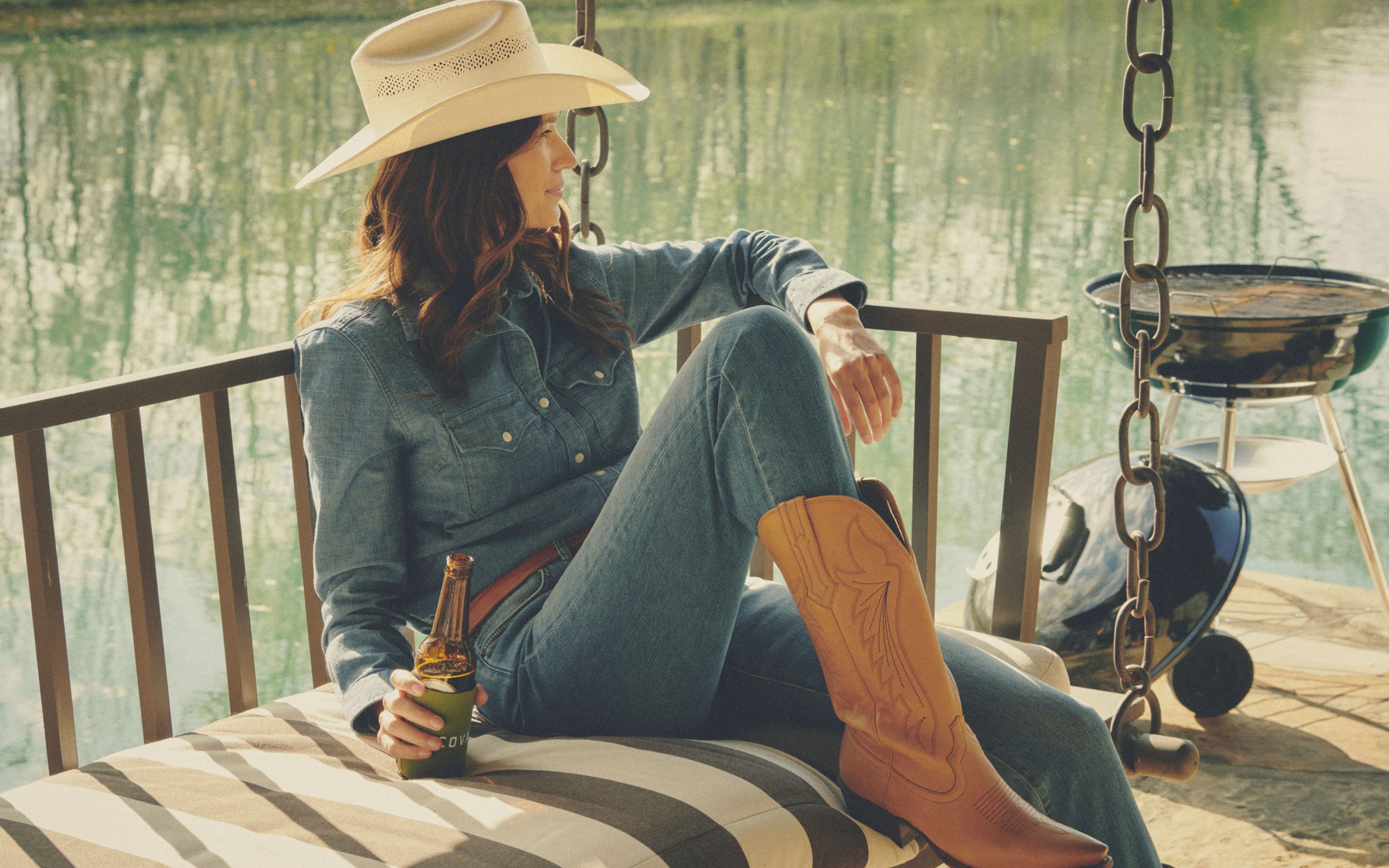 Woman in cowboy hat, jeans, and boots drinking a beer on a porch swing by a lake