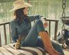 Woman in cowboy hat, jeans, and boots drinking a beer on a porch swing by a lake