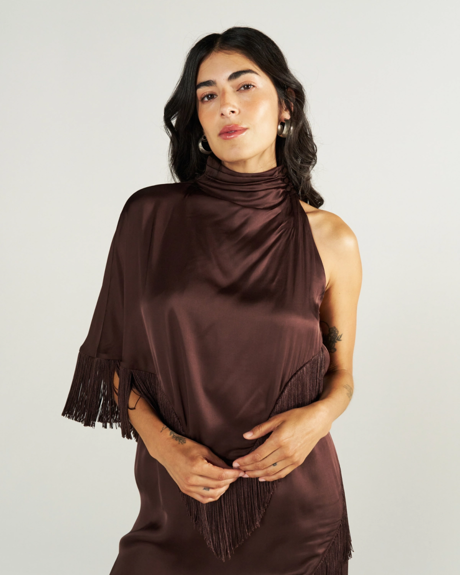 Woman with long dark hair wearing a shiny, dark brown, asymmetrical dress with fringe detail, posing against a plain light background.