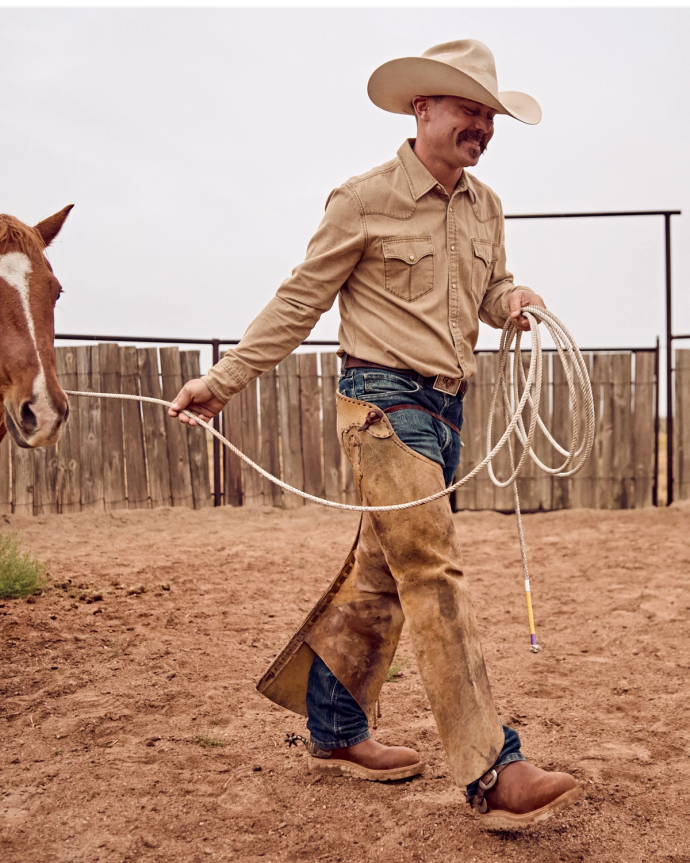 Man in a cowboy hat and chaps leading a horse with a rope in a fenced dirt area.