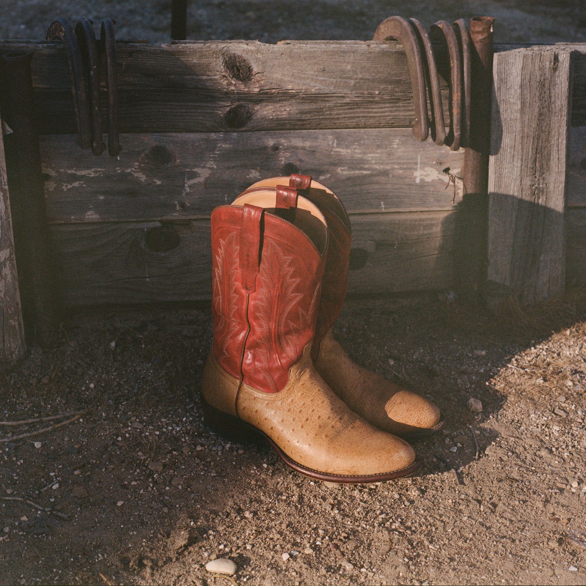 A pair of tan cowboy boots with red tops resting on dirt in front of a weathered wooden fence with horseshoes hanging on it.