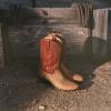 A pair of tan cowboy boots with red tops resting on dirt in front of a weathered wooden fence with horseshoes hanging on it.