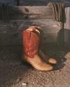 A pair of tan cowboy boots with red tops resting on dirt in front of a weathered wooden fence with horseshoes hanging on it.