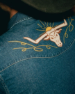 Close-up of a denim shirt with an embroidered longhorn skull and sun design on the back, worn by a person in a dark hat.