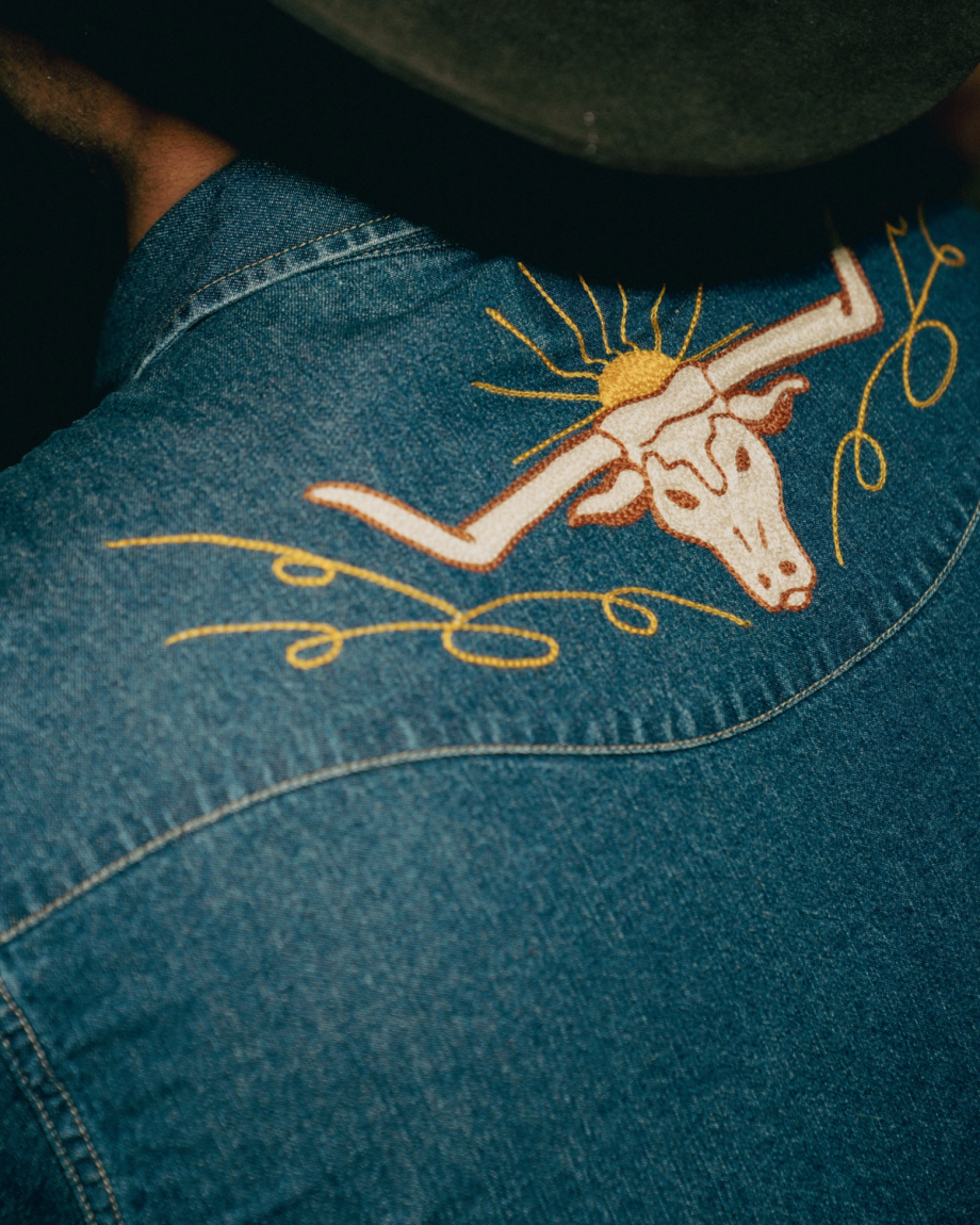 Close-up of a denim shirt with an embroidered longhorn skull and sun design on the back, worn by a person in a dark hat.
