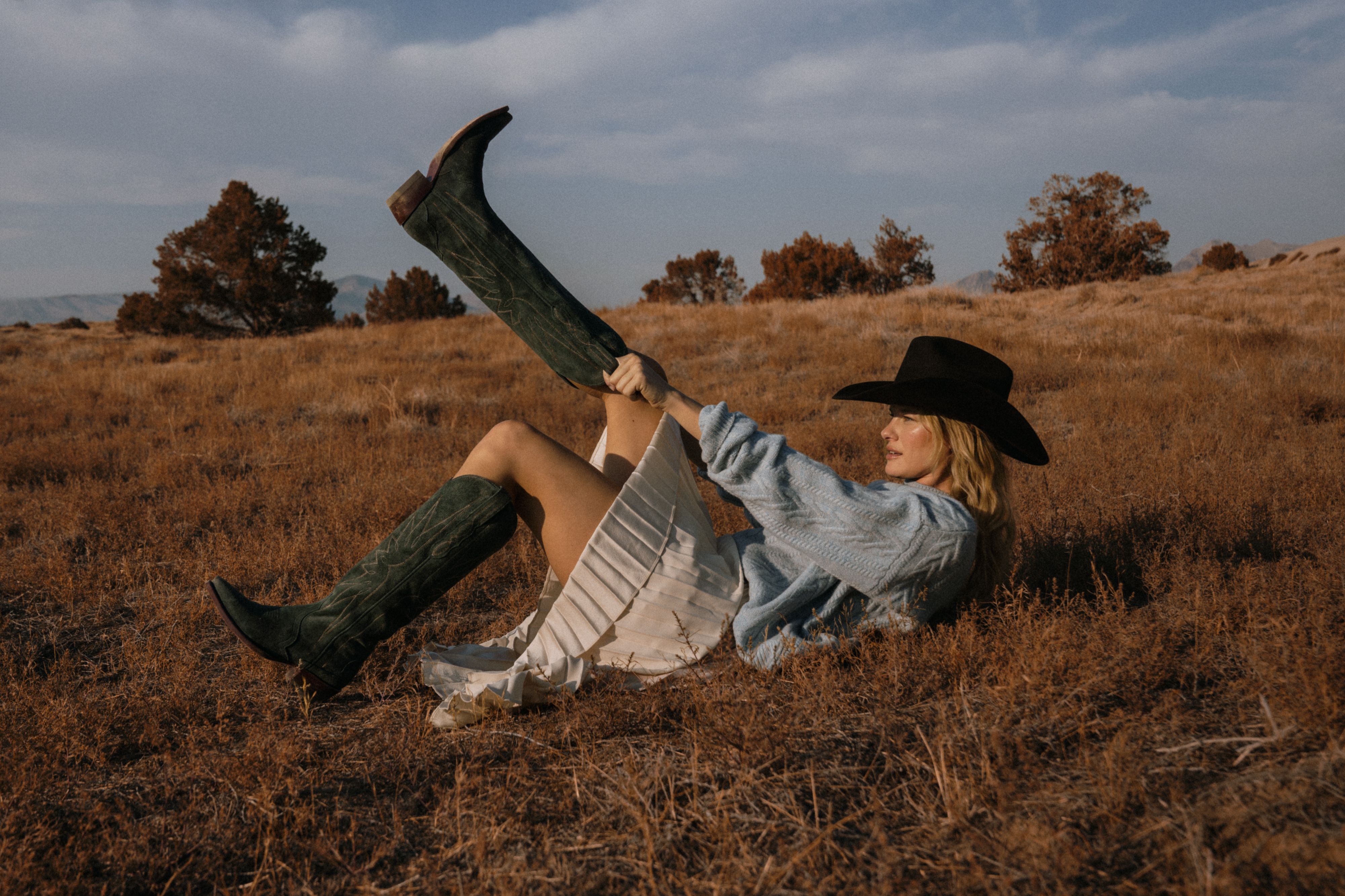 A person in a black cowboy hat, blue sweater, and white skirt lying in a field while pulling on a dark green cowboy boot.