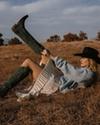 A person in a black cowboy hat, blue sweater, and white skirt lying in a field while pulling on a dark green cowboy boot.