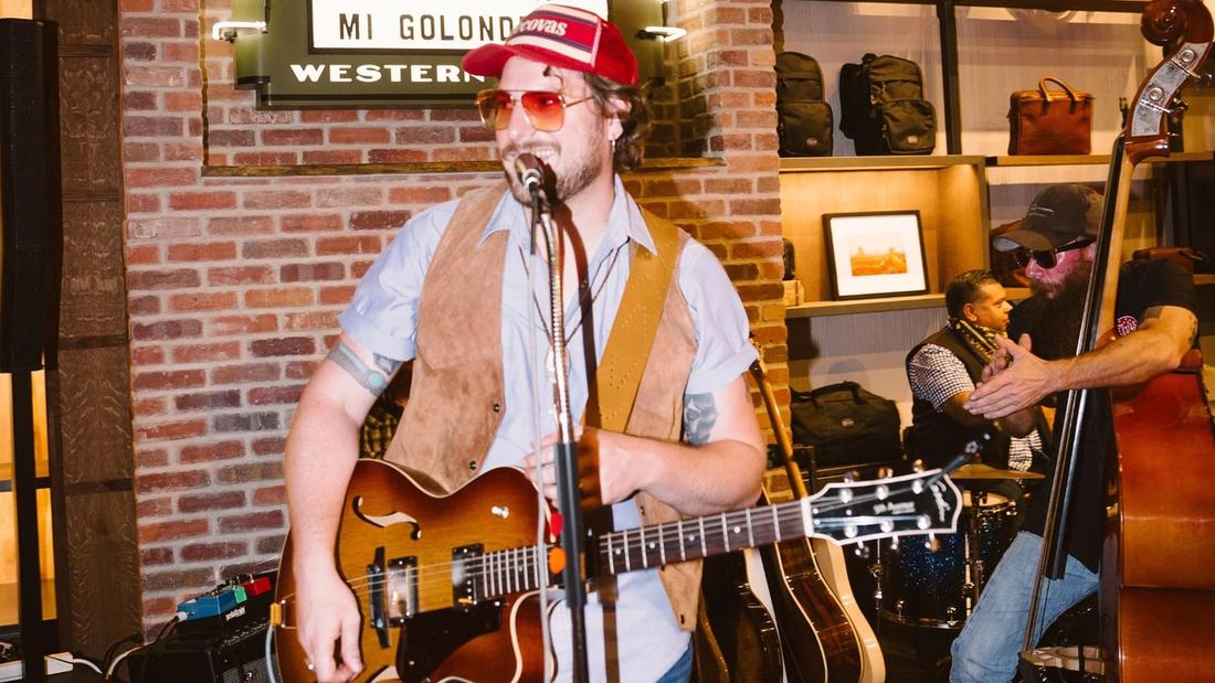 A musician wearing a red cap and sunglasses plays a guitar and sings into a microphone while another musician tunes a double bass in the background. They are in a brick-walled space with various items displayed on shelves.