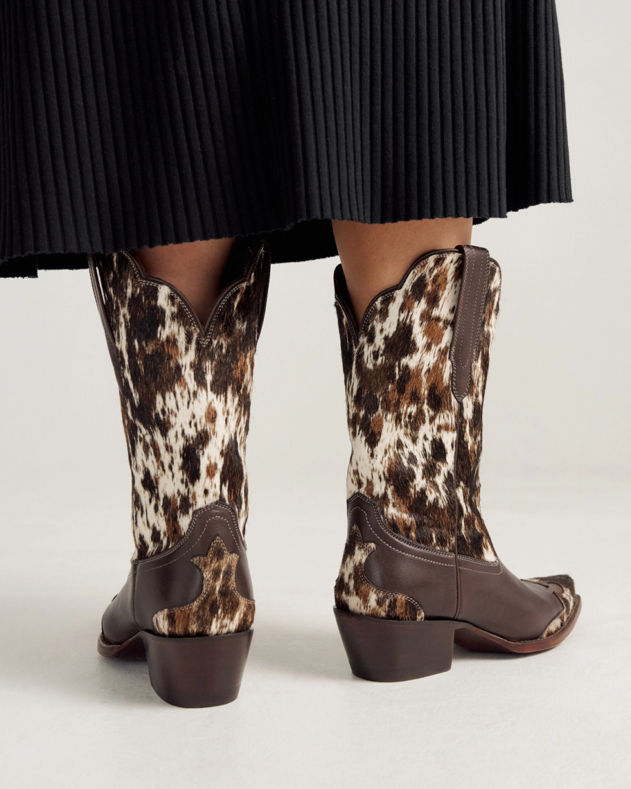 A person wearing The Sadie Hair on Hide / Cowhide cowboy boots stands on a light-colored floor, with a pleated black skirt visible above the boots.
