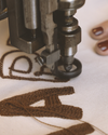 Close-up of an embroidery machine stitching brown thread into fabric, with a partial view of a person’s hand with red nail polish beside the machine.
