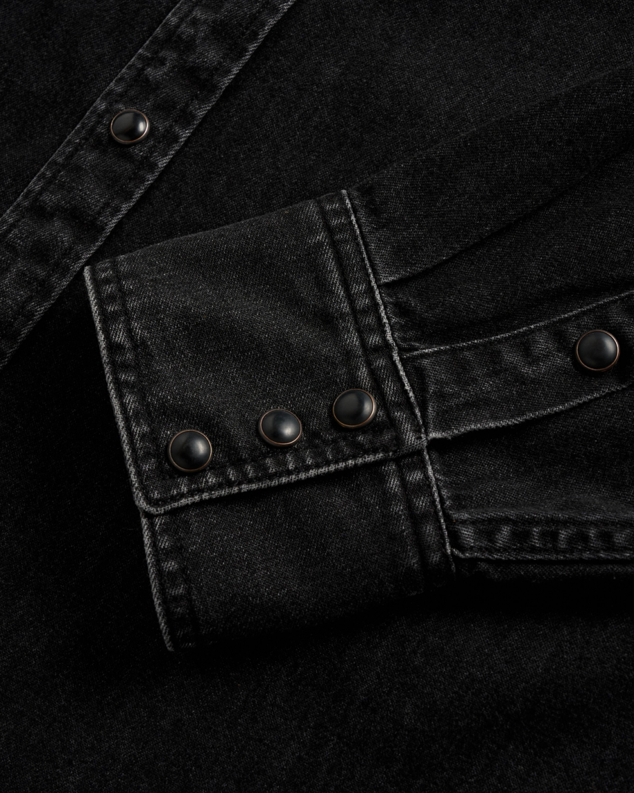 Close-up of a black denim shirt cuff with three black snap buttons and visible white stitching.