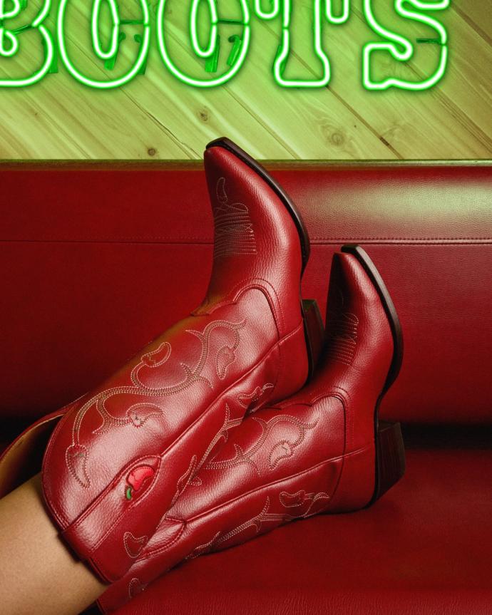 Person wearing red cowboy boots reclines on a red couch; a neon green sign reading "BOOTS" is partially visible in the background.