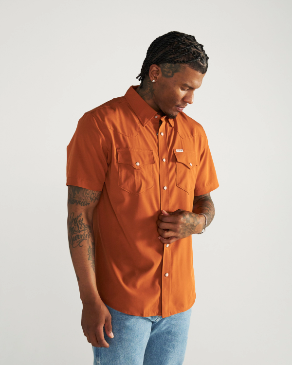 Man wearing an orange short-sleeve button-up shirt with two chest pockets and blue jeans, standing against a plain white background.
