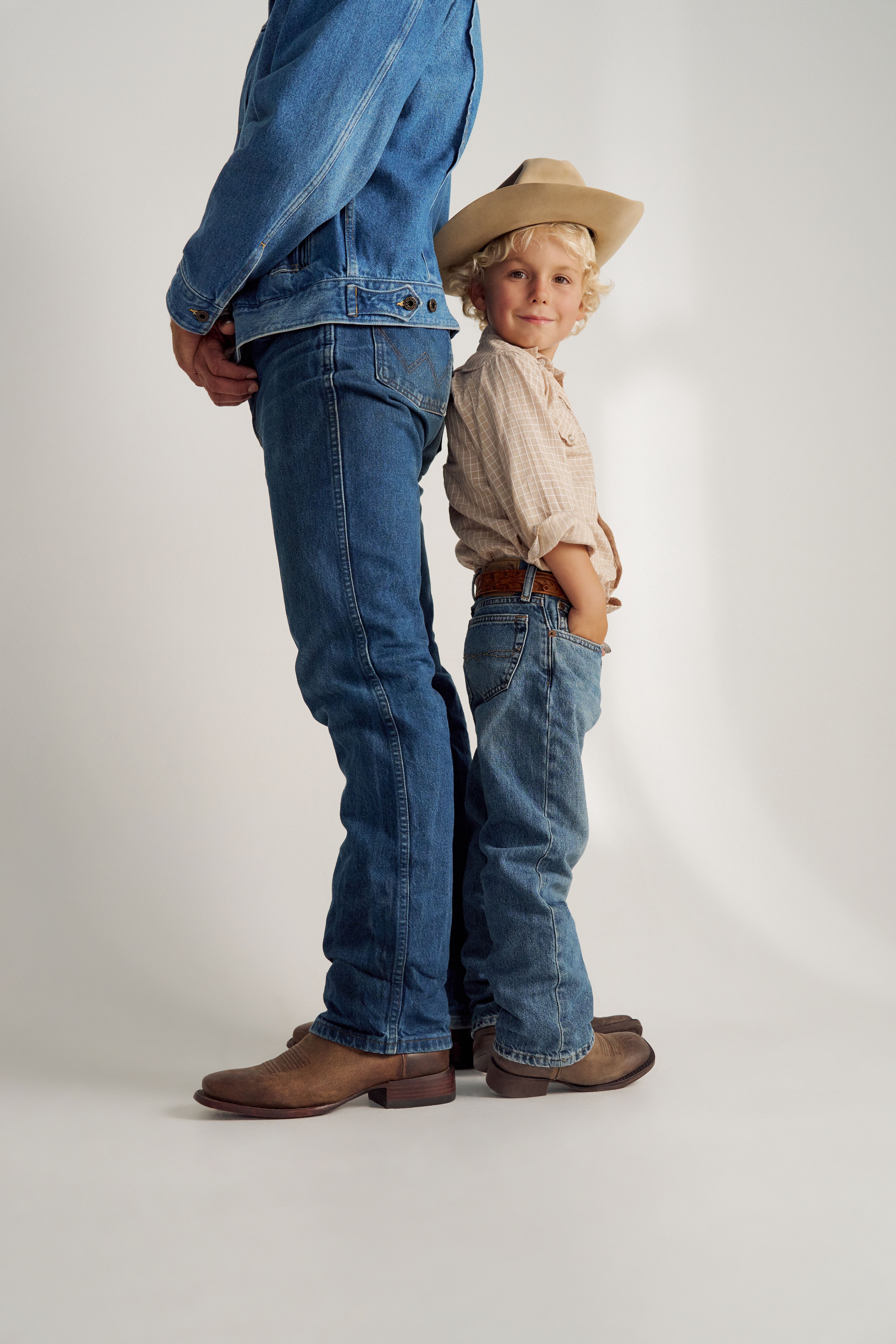 Two people wearing blue jeans and brown leather shoes stand close together, facing each other on a neutral background.