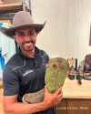 A man wearing a cowboy hat and a black polo shirt is holding a green boot. He is standing in front of a wooden table with various items on it.