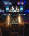 Two cowboys on stage with large screen and pyrotechnics at a rodeo event. Audience members are visible in the foreground.