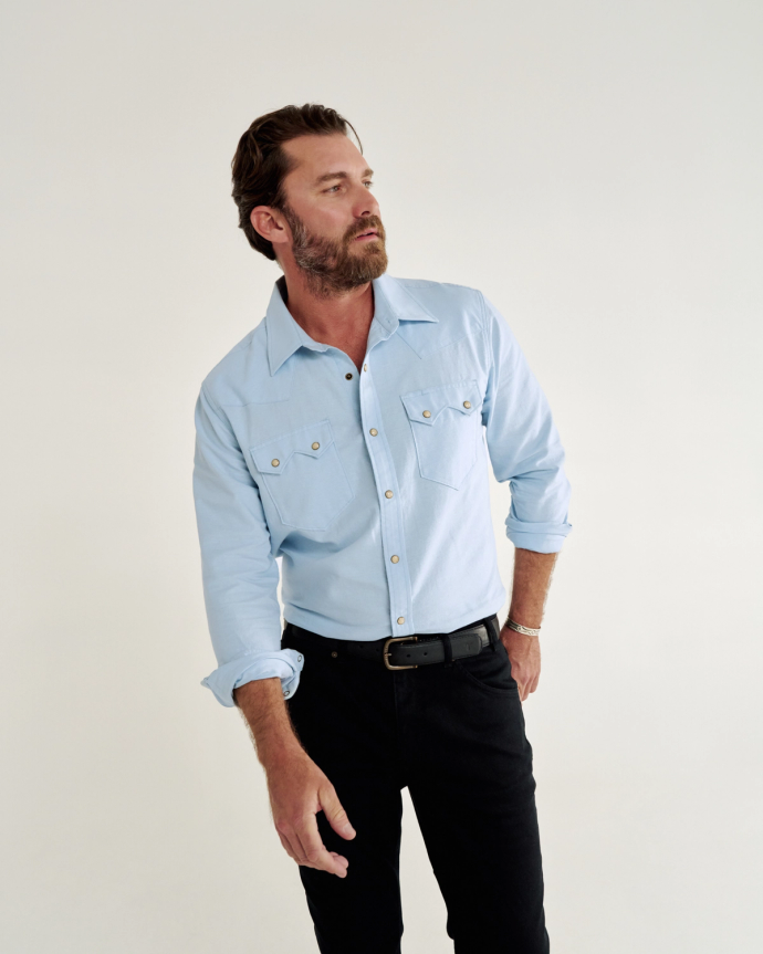 A man with a beard wearing a light blue button-up shirt and black pants, standing against a plain white background.