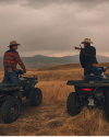 Two people in cowboy hats on ATVs pause in a field. One gestures towards the distant hills. The sky is overcast and the landscape is expansive and rugged.