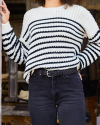 A person wearing a black and white striped sweater and black jeans stands with one hand resting on a vertical surface.