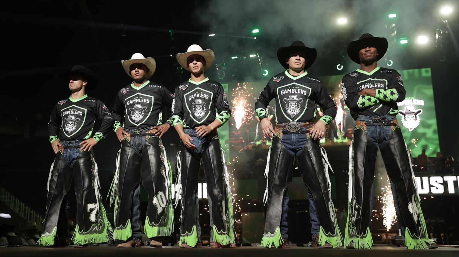 Five men in matching black and green cowboy outfits stand in a line, wearing helmets and chaps, with fireworks and green lights in the background.