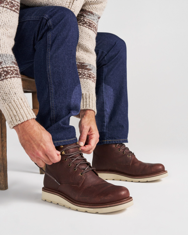 The Knox Lace Up in Cognac Cowhide. A person wearing dark blue jeans and a patterned sweater ties the laces of a cognac cowhide boot with a thick white sole.