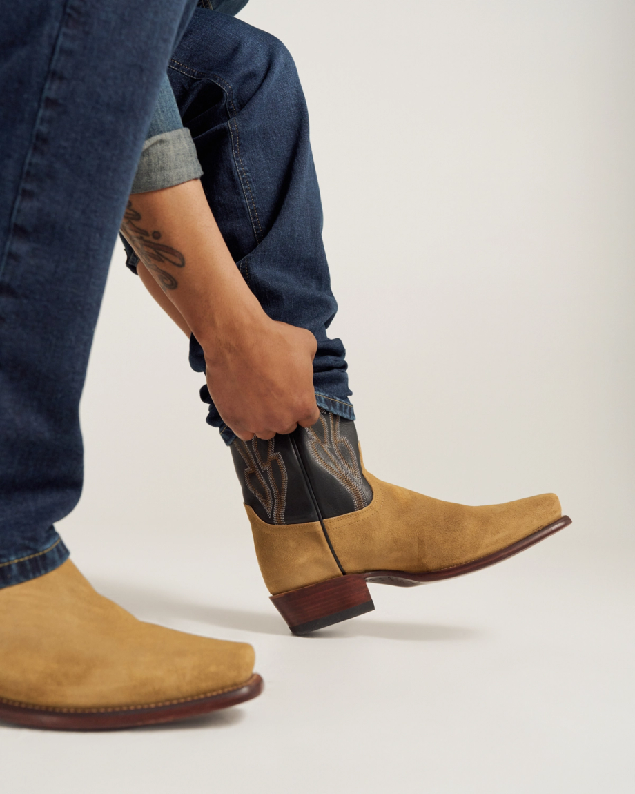 Person in blue jeans pulling on tan cowboy boots with dark stitching detail; visible tattoo on forearm.
