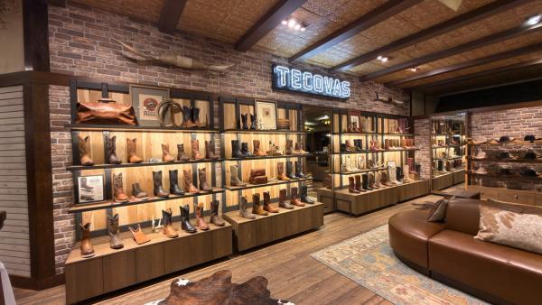 Interior of a Tecovas retail store featuring shelves of leather cowboy boots, a brick wall, and a neon sign with the brand name.