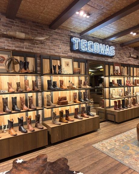 Interior of a Tecovas retail store featuring shelves of leather cowboy boots, a brick wall, and a neon sign with the brand name.