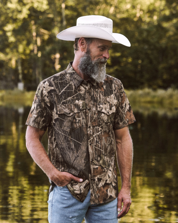 Man with a gray beard wearing a white cowboy hat, camouflage shirt, and blue jeans stands outdoors near water, with trees in the background.
