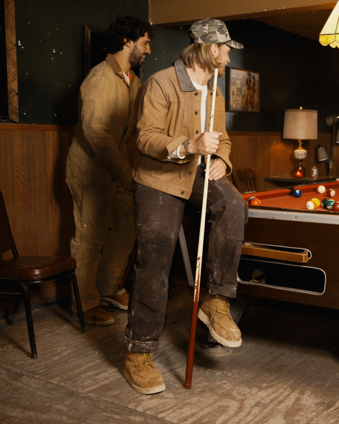 Two people in casual workwear play pool indoors. One holds a pool cue and leans on the table, while the other stands nearby. Pool balls are visible on the table.