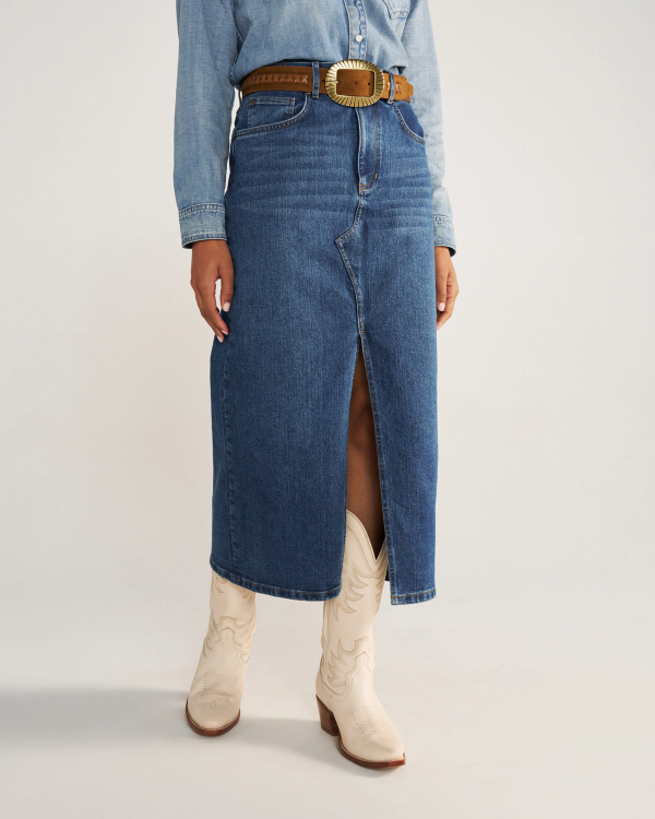 A person wearing a blue denim skirt with a front slit, a brown belt, a denim shirt, and white cowboy boots stands against a plain background.