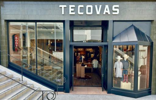 Exterior view of a Tecovas store with glass doors and windows displaying boots and clothing. Stairs are on the left side, and a neon "BOOTS" sign is visible inside.