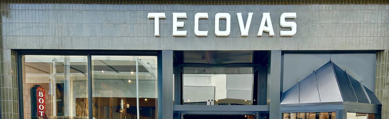 Exterior view of a Tecovas store with glass doors and windows displaying boots and clothing. Stairs are on the left side, and a neon "BOOTS" sign is visible inside.