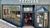 Exterior view of a Tecovas store with glass doors and windows displaying boots and clothing. Stairs are on the left side, and a neon "BOOTS" sign is visible inside.