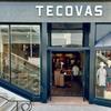 Exterior view of a Tecovas store with glass doors and windows displaying boots and clothing. Stairs are on the left side, and a neon "BOOTS" sign is visible inside.